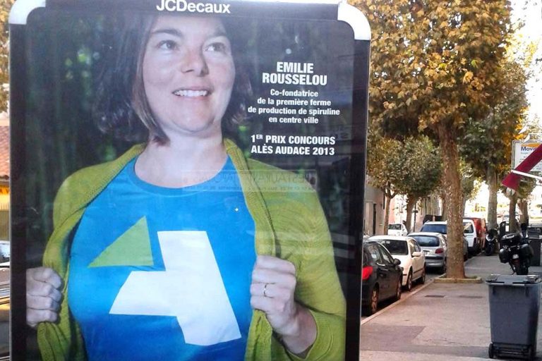 Femme souriante portant un t-shirt bleu avec un logo sur une rue bordée d'arbres.